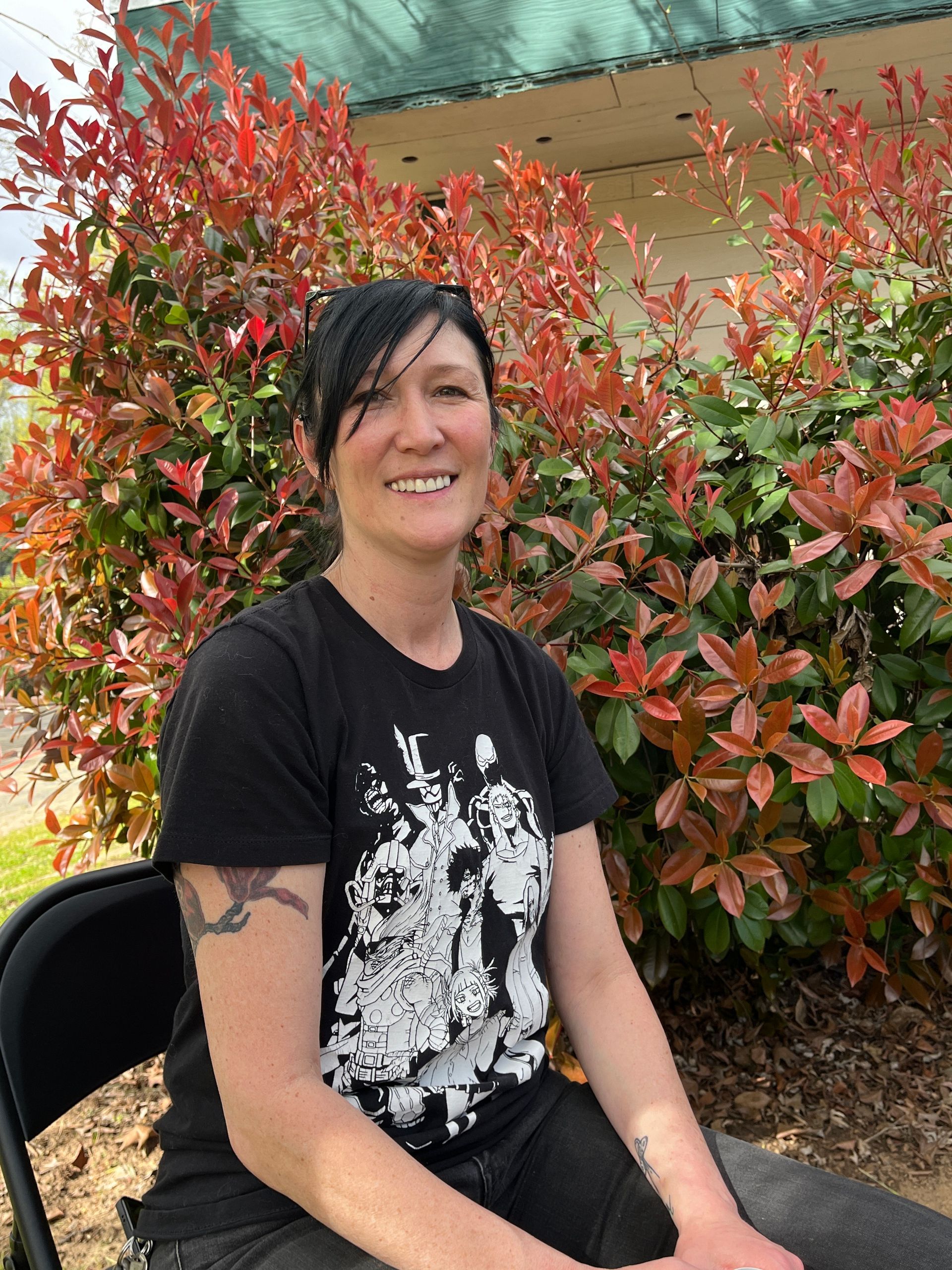 Woman with dark hair, black t-shirt, smiling, sits outside with red-leafed shrubs behind her.