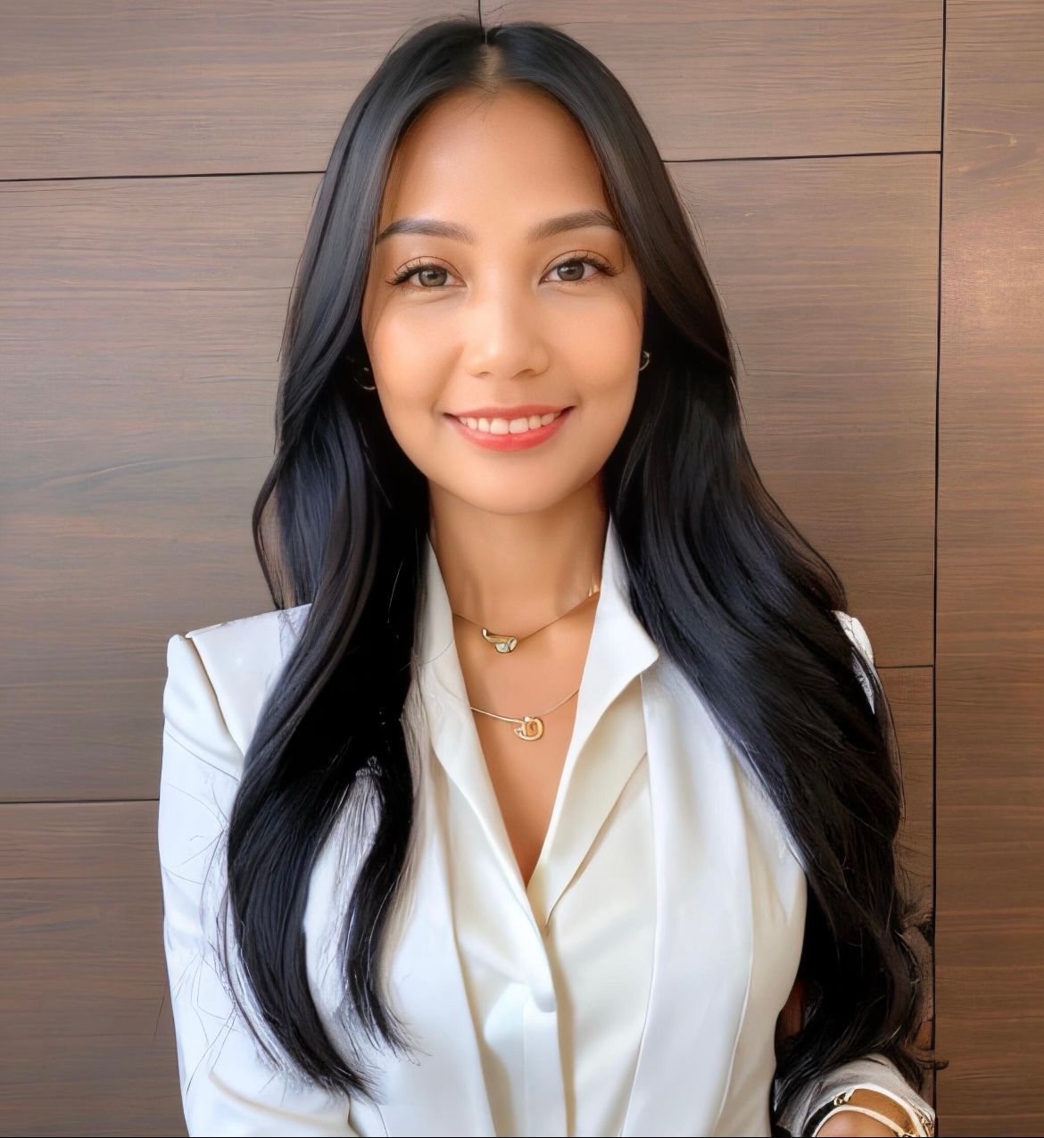 Woman in white blazer smiling, long black hair, gold necklace, against a brown wall.