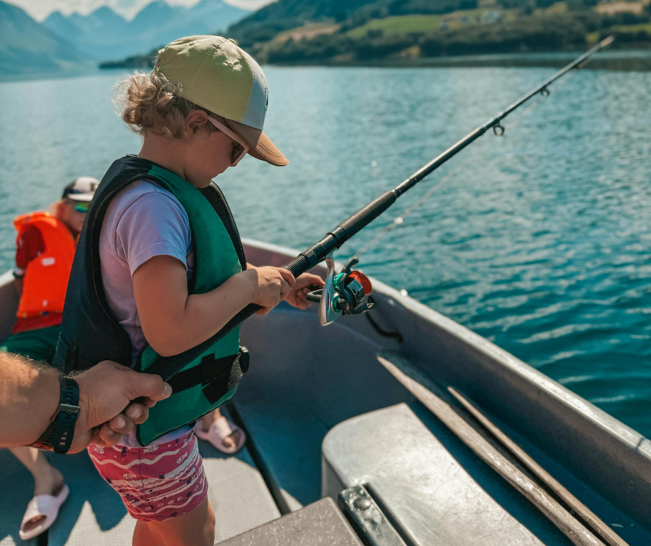 Child fishing from a boat, wearing a life vest and hat, holding a fishing rod, with mountains in the background.