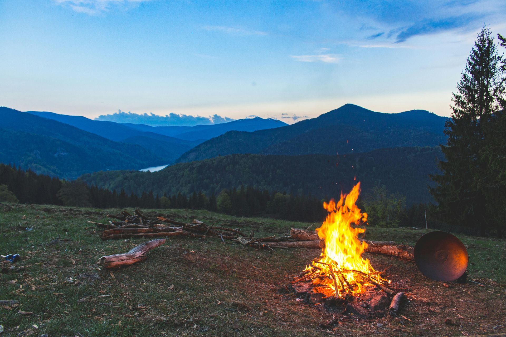 Campfire in mountain landscape at dusk; burning flames, trees, and layered blue-green hills.