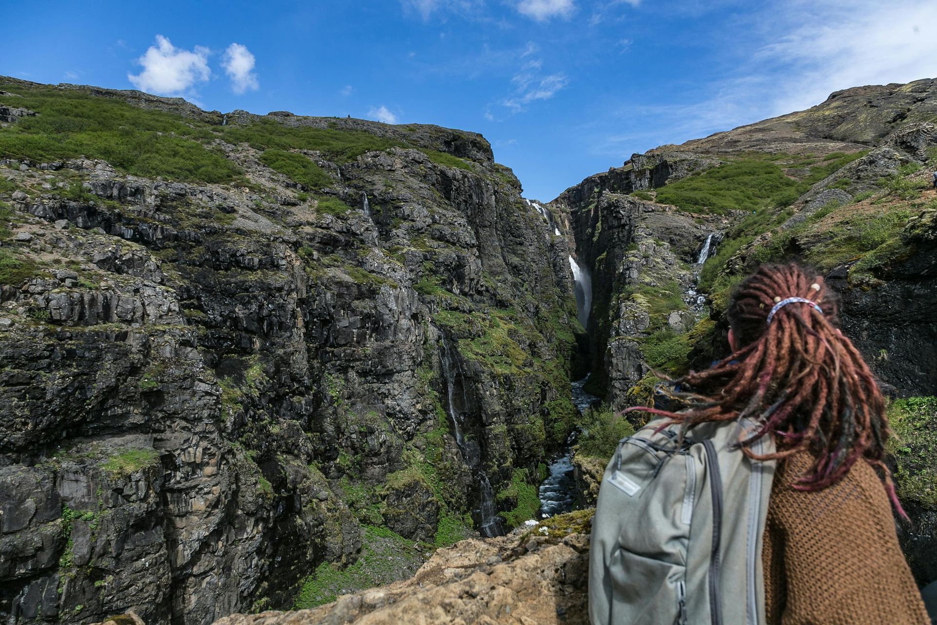 Person with dreadlocks, backpack overlooking a waterfall and rocky canyon under a blue sky.