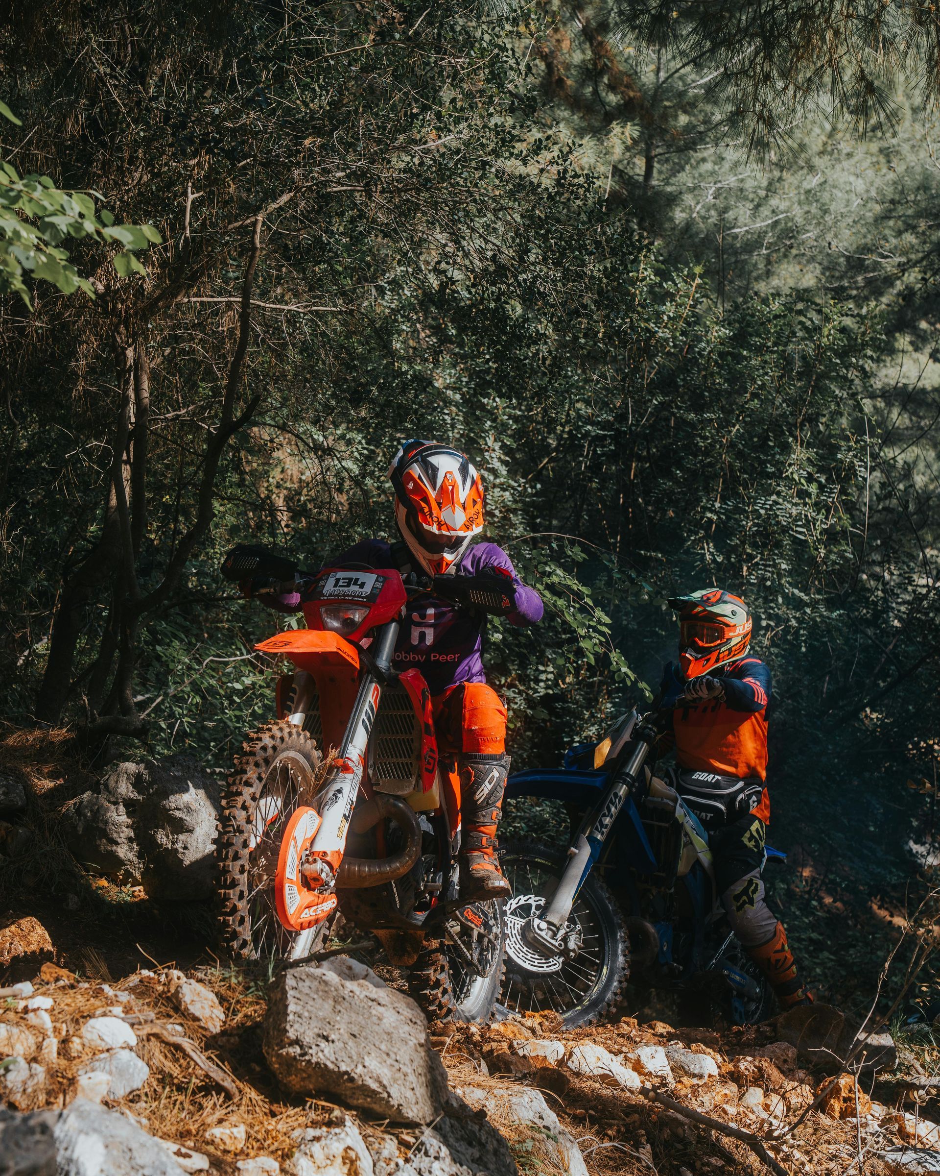 Two people on dirt bikes riding through a wooded area. Orange and purple gear.