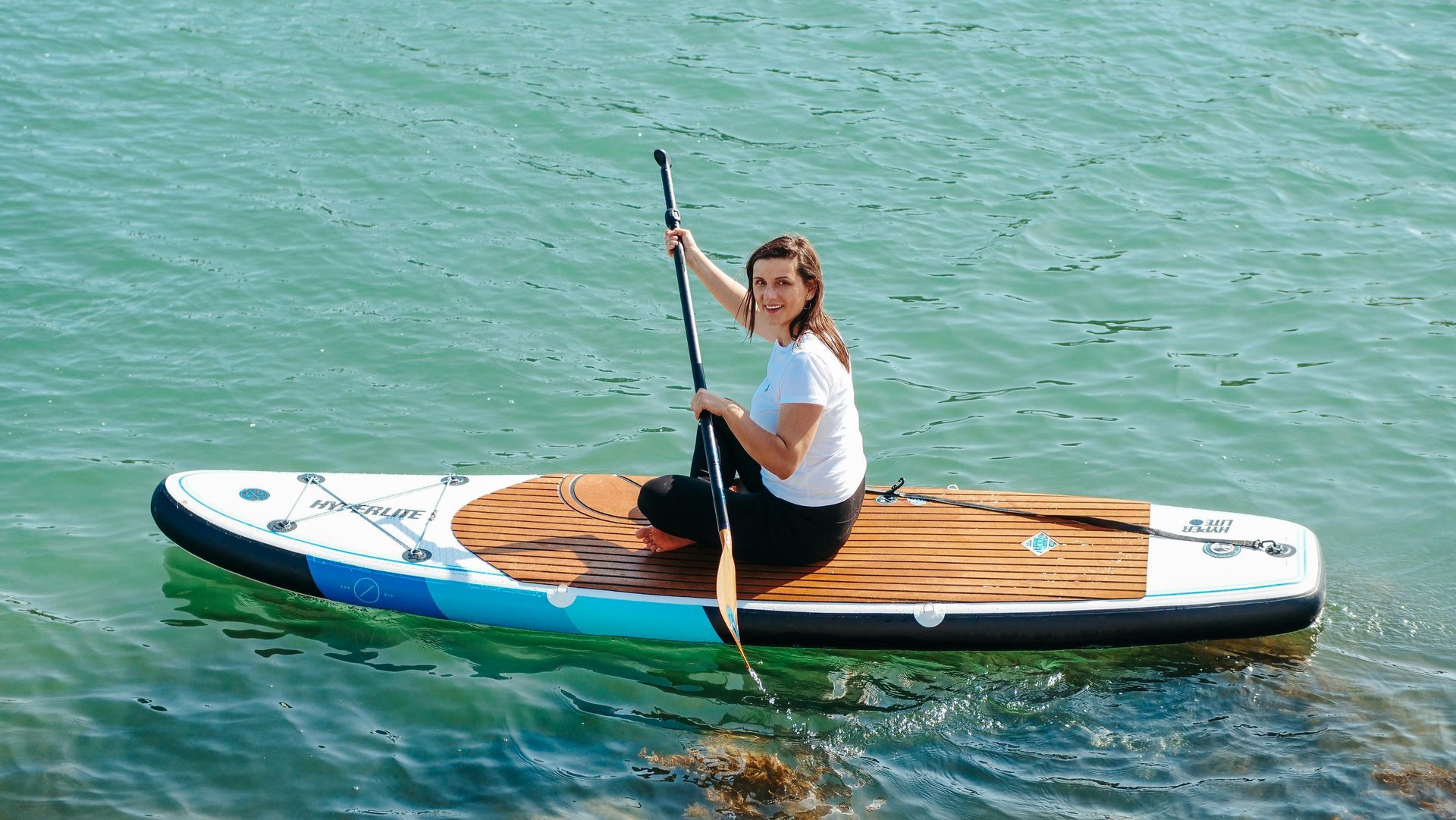 Woman on a paddleboard smiles, holding paddle. She is in the water on a sunny day.