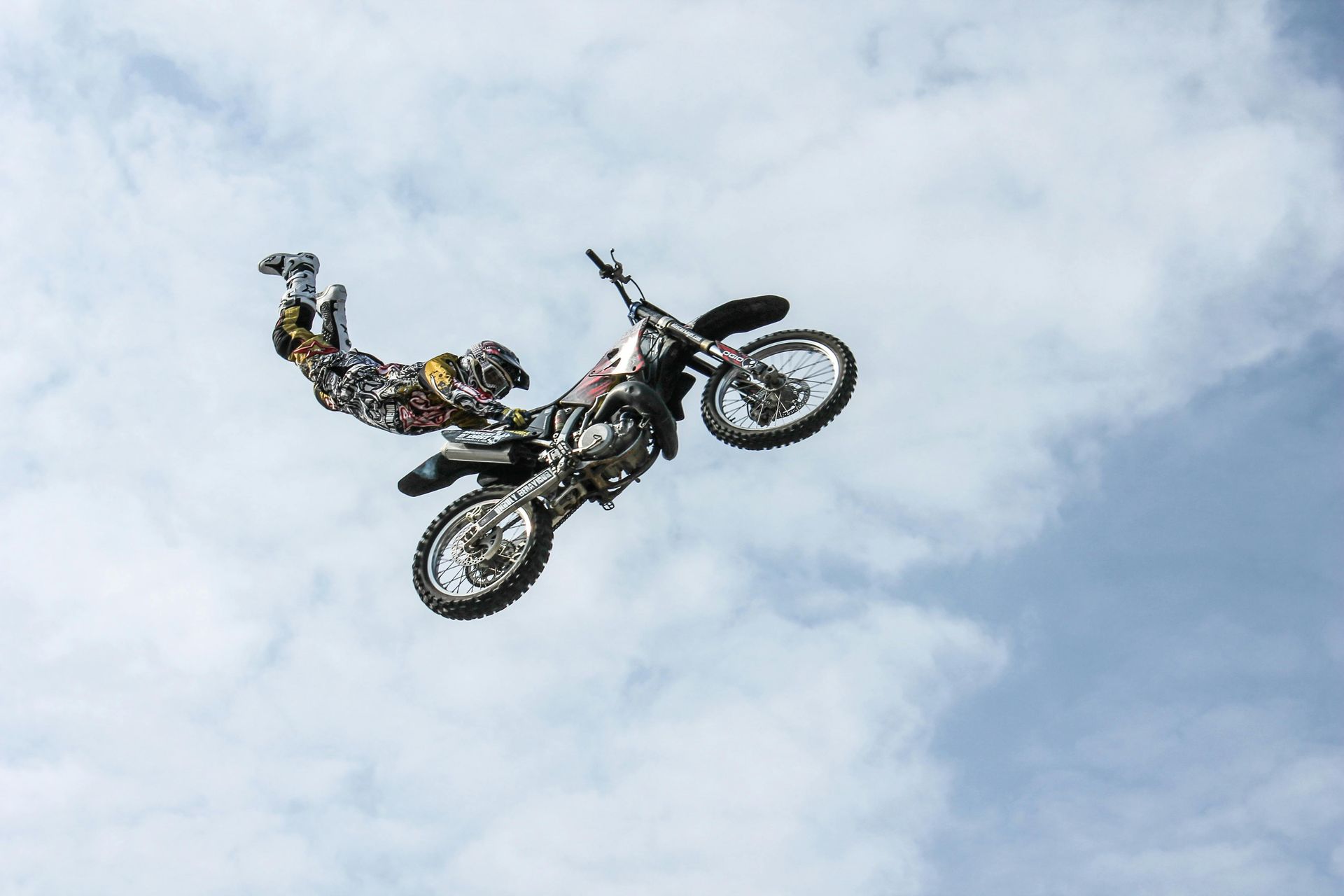 Motorcycle rider in mid-air doing a stunt, against a partly cloudy sky.