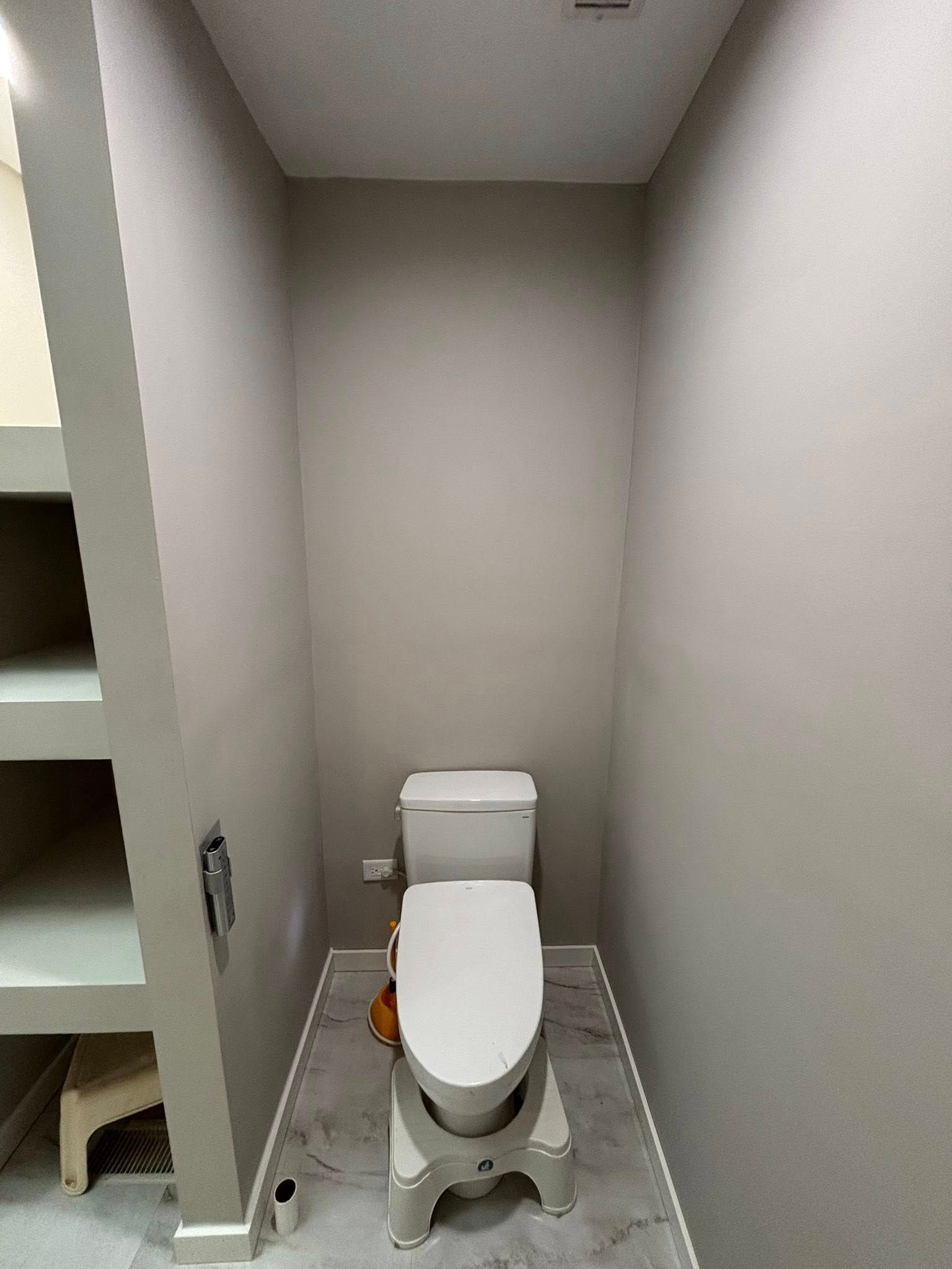 Small, narrow bathroom with a white toilet, gray walls, and a small step stool.