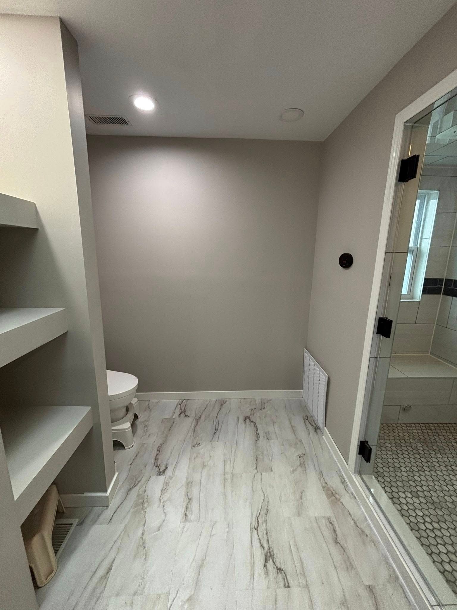 Bathroom with built-in shelves on the left, toilet in the middle, shower on the right, and wood-look flooring.