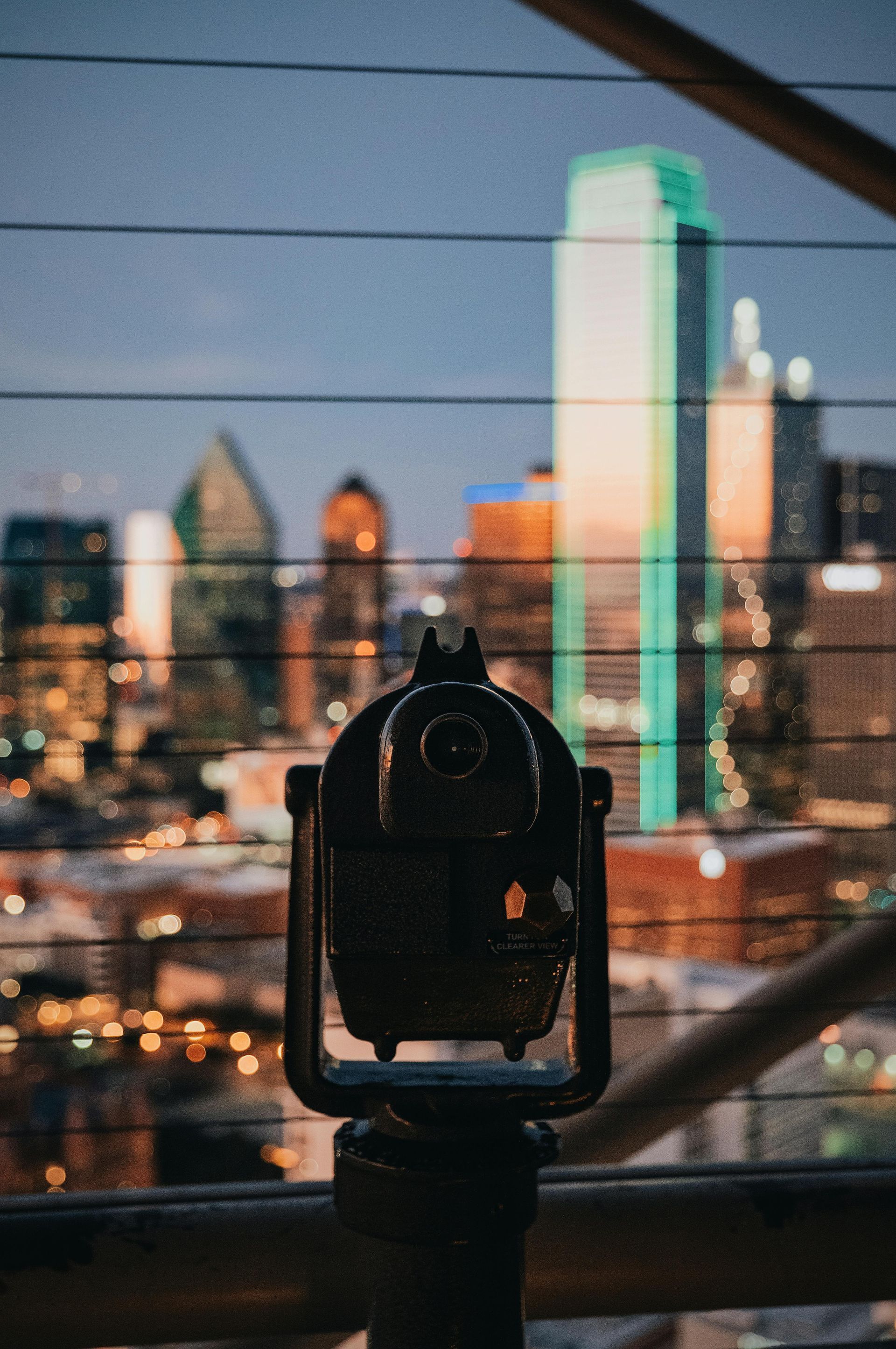 City skyline viewed through a black viewer. Buildings reflect colorful sunset hues with a grid of wires in the foreground.