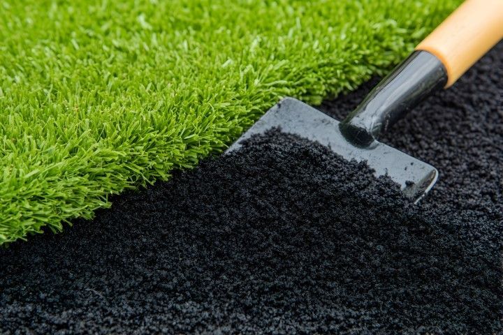 A spade partially lifts a synthetic turf, revealing black crumb rubber underneath