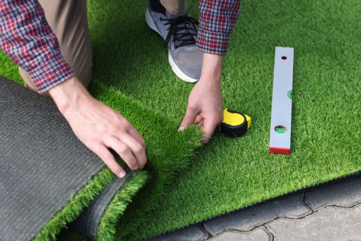 Man lays artificial grass on patio, using a tape measure and level. 