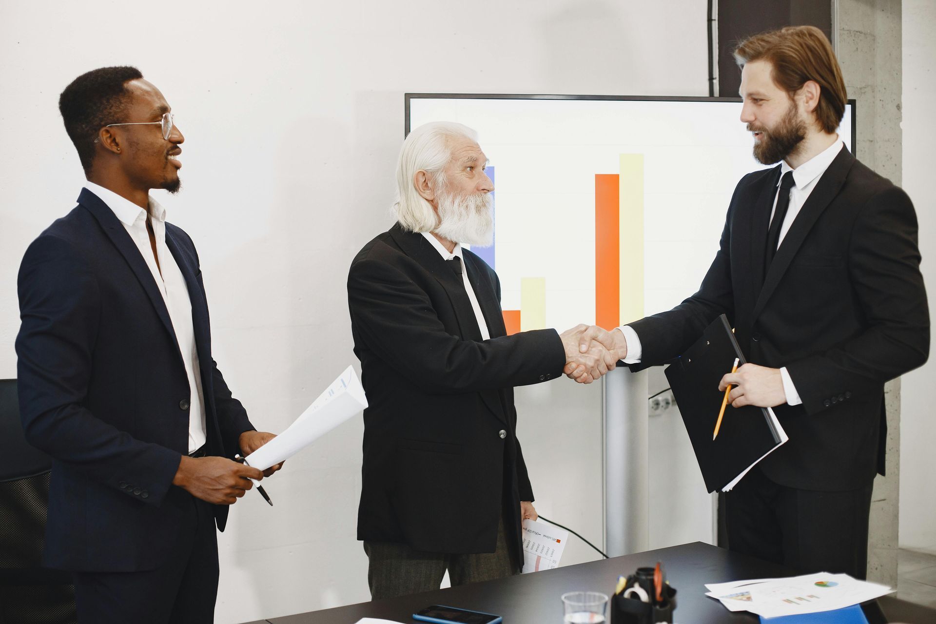 Three people in business attire shake hands in an office with a bar graph displayed on a screen in the background.