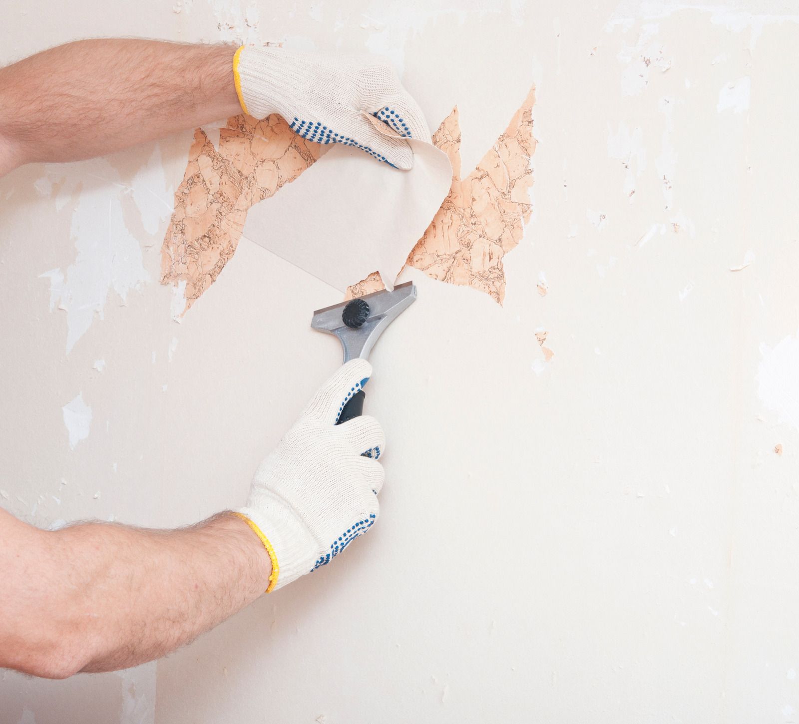Hands wearing gloves peeling wallpaper with a scraper tool; beige wall.