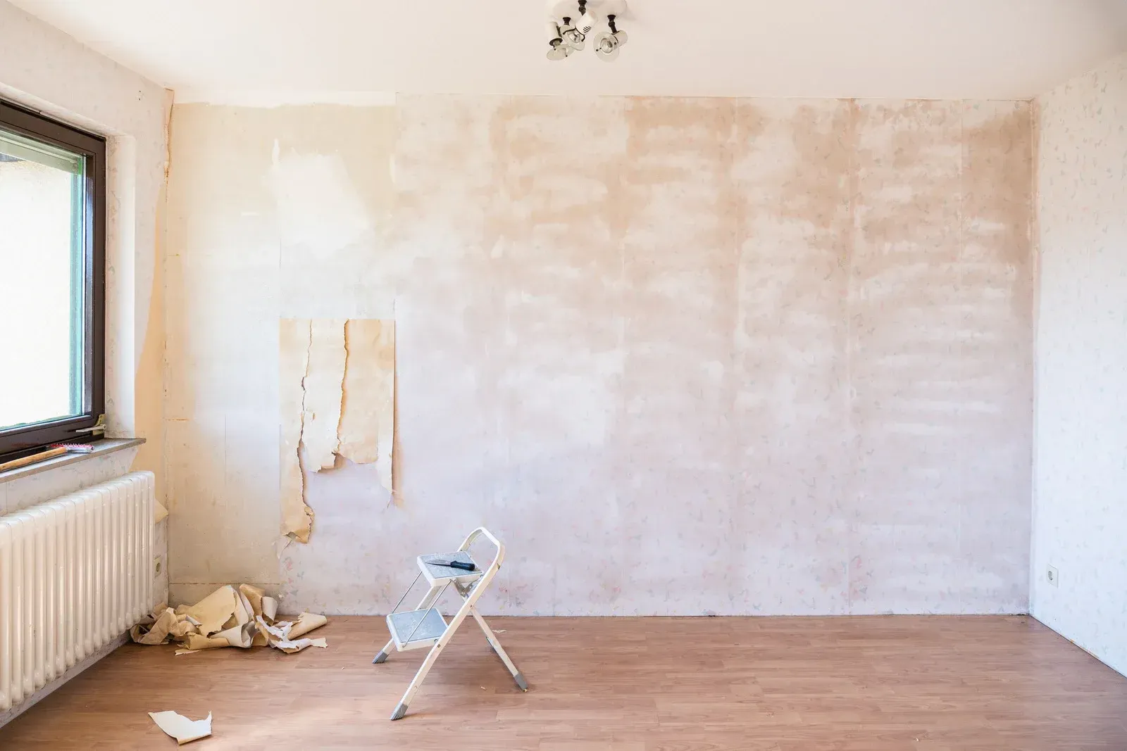 Room undergoing renovation; wallpaper being removed from beige wall. Small ladder and debris on wood floor.