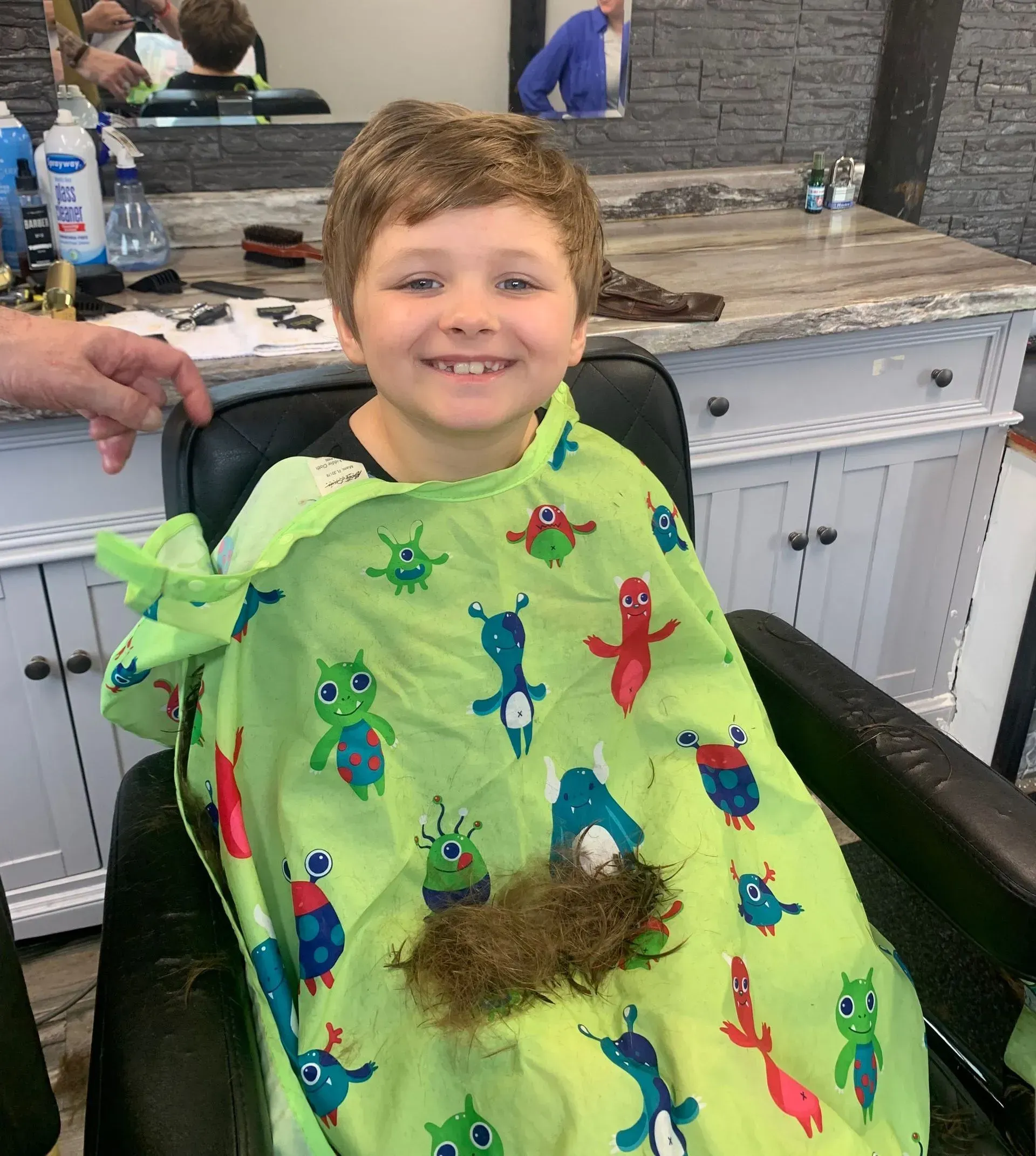 A young boy is sitting in a chair with a green cape on his head.