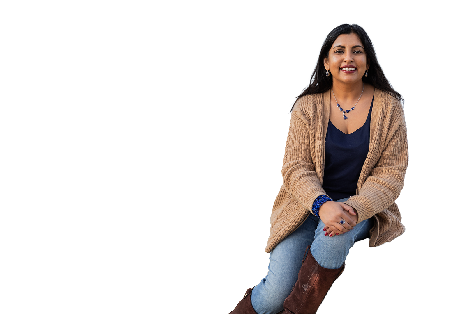 Woman seated on a stool against a white background, smiling in a tan cardigan and blue top.