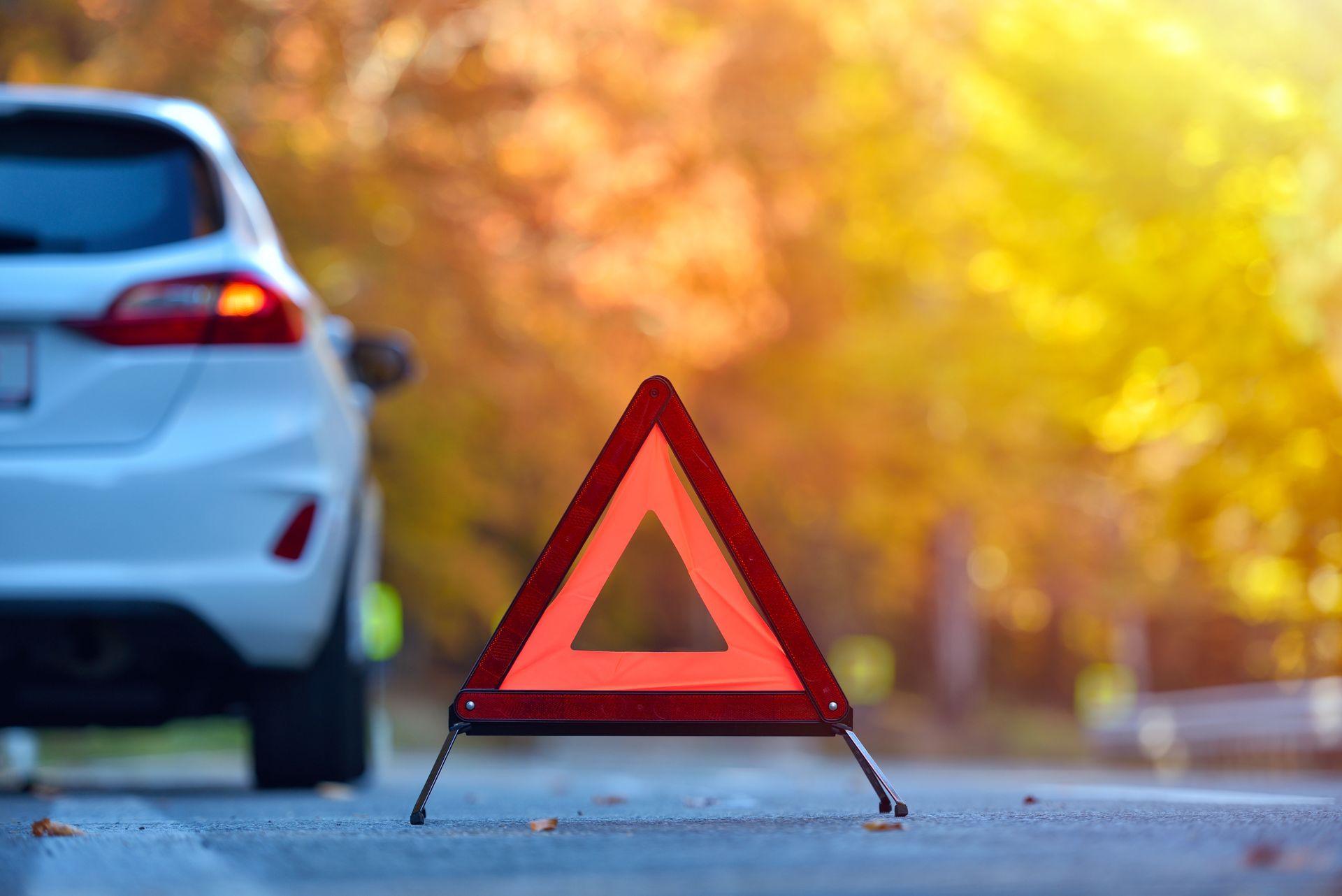White car with hazard triangle on roadside, autumn trees in background.