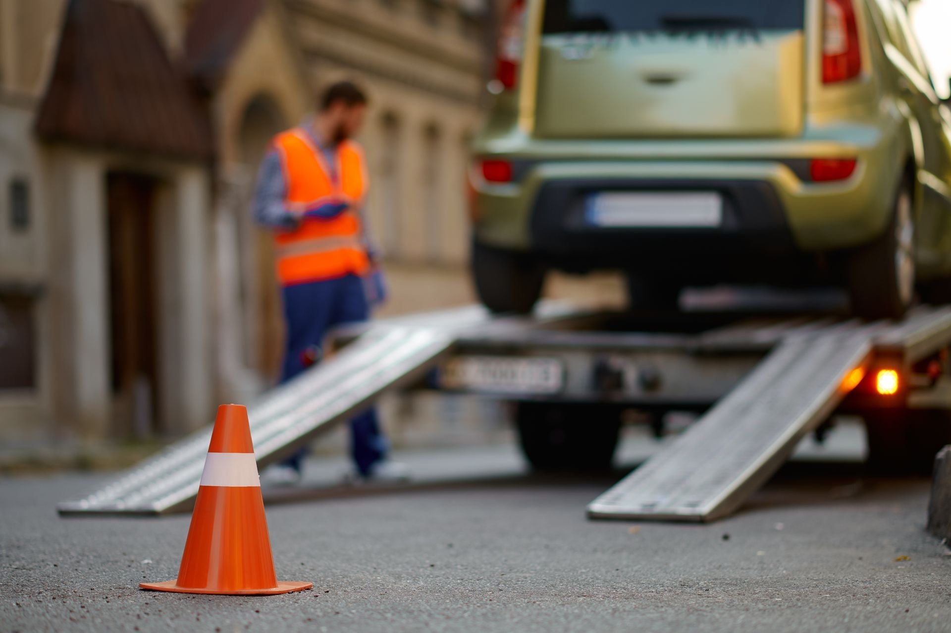 Orange traffic cone in foreground; tow truck loading a green car. A worker in an orange vest looks on.