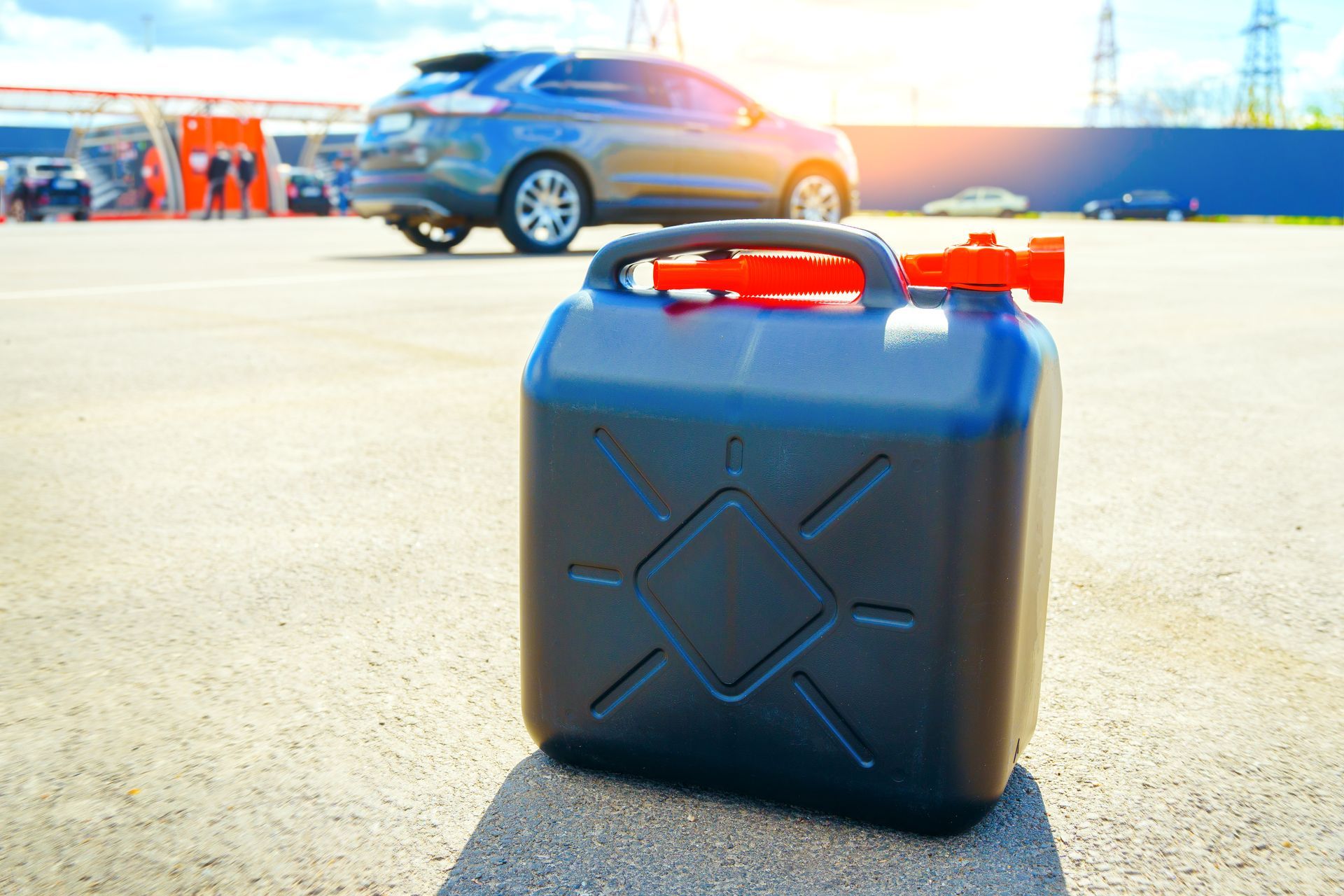 Black gas can with red spout, in a parking lot with a car and car wash in the background.