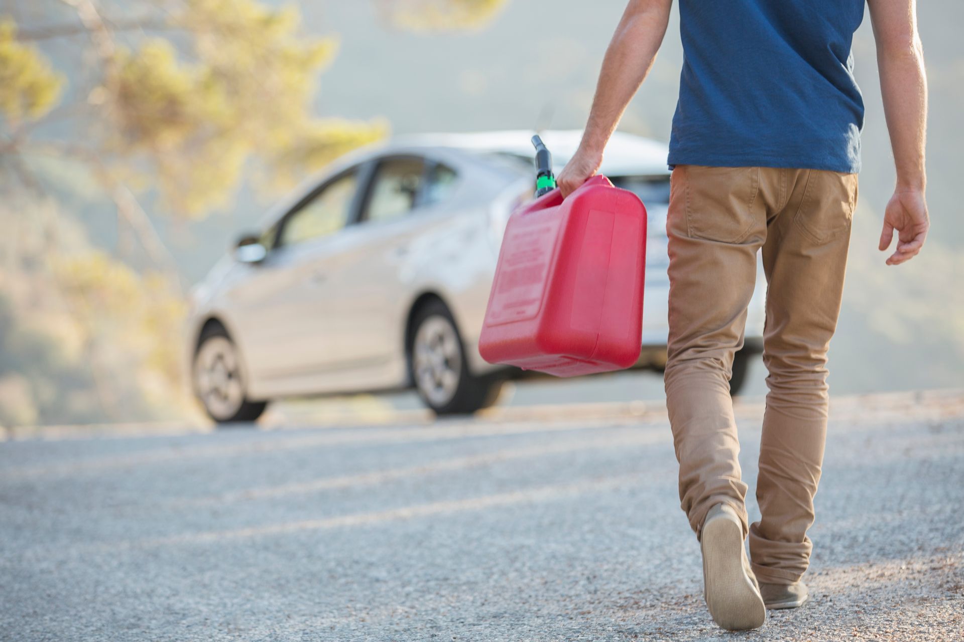 Person carrying a red gas can towards a car on a road.