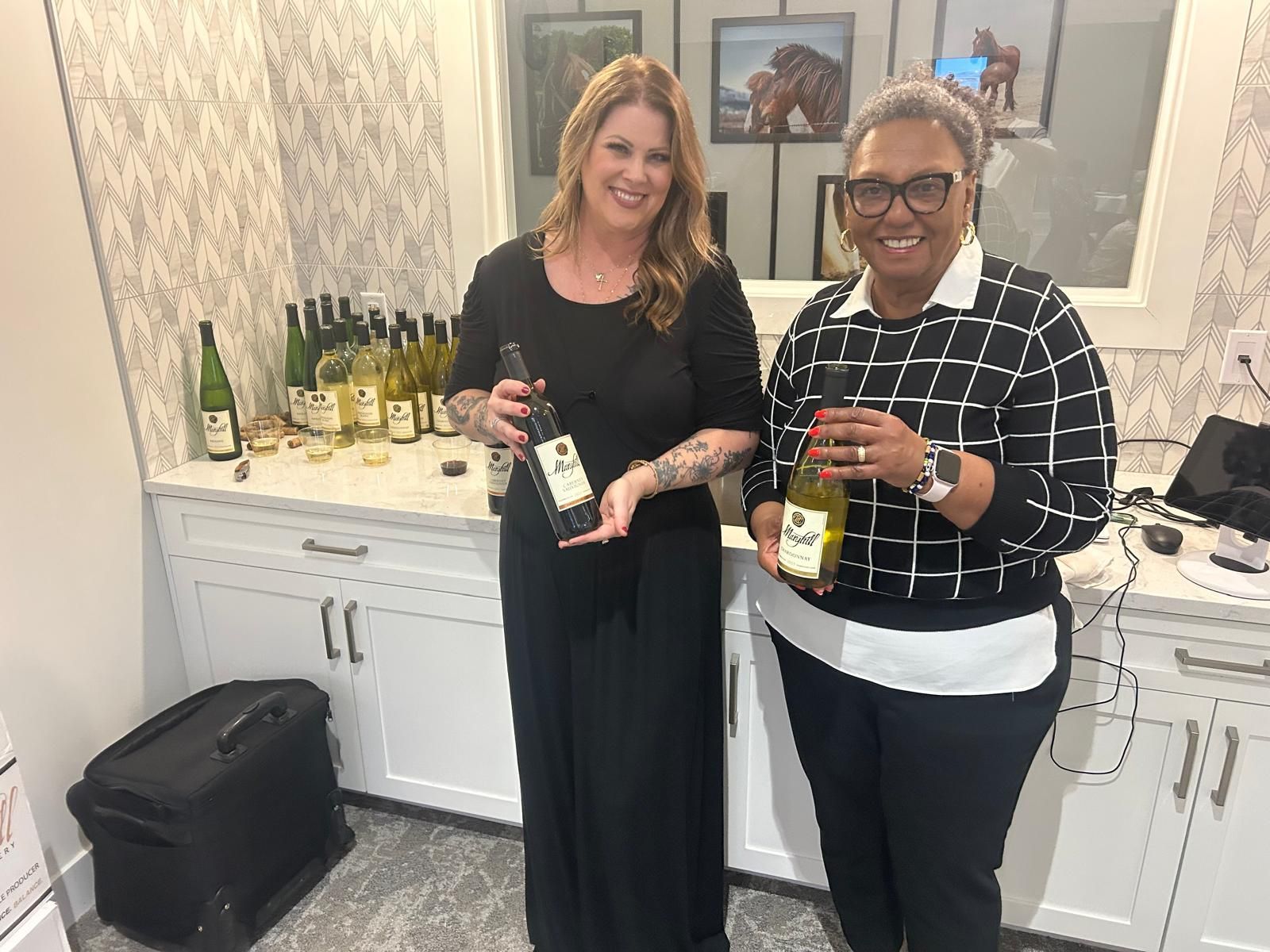 Two women are standing next to each other in a kitchen holding bottles of wine.