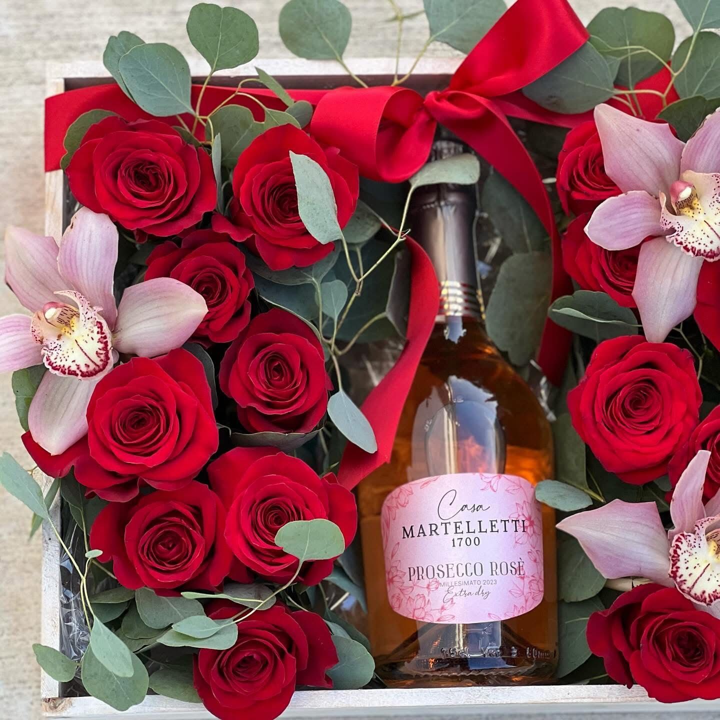 A bottle of wine sits in a box surrounded by red roses