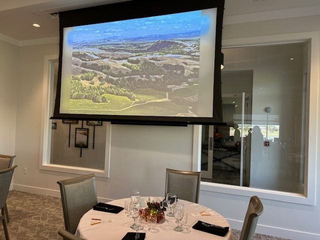 A large screen is hanging from the ceiling in a room with a table and chairs.