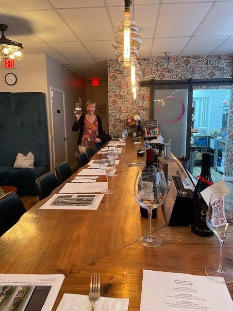 A woman is standing behind a long wooden table holding a glass of wine.