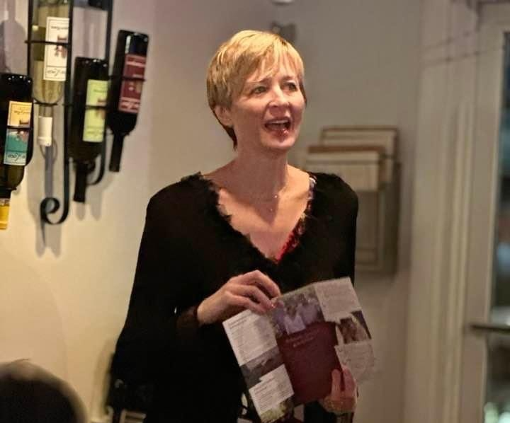 A woman is holding a pamphlet in front of a wall of wine bottles.
