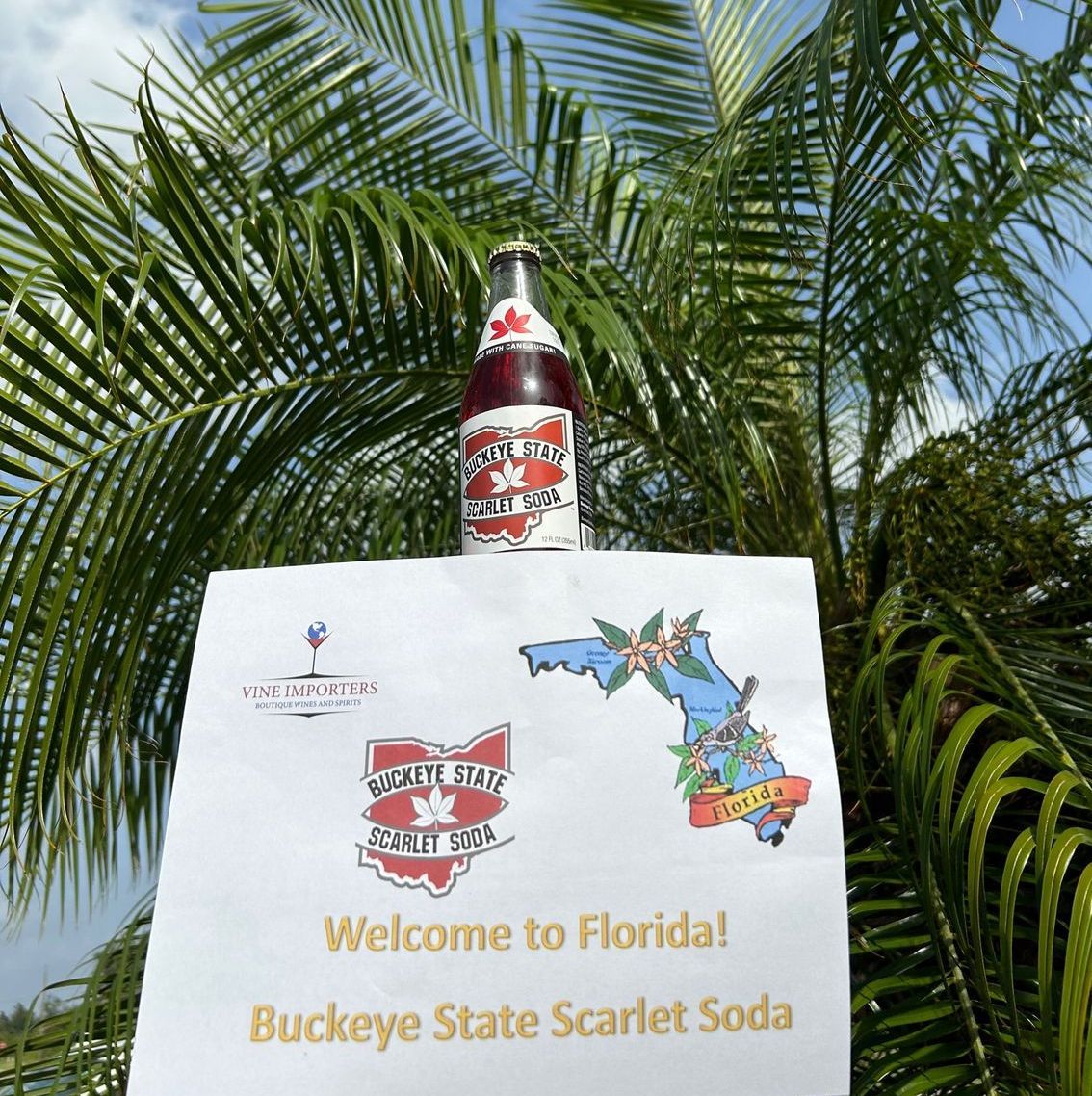 A bottle of buckeye state scarlet soda sits on top of a welcome to florida sign