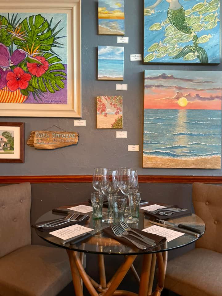 There are many paintings on the wall behind the table and chairs.