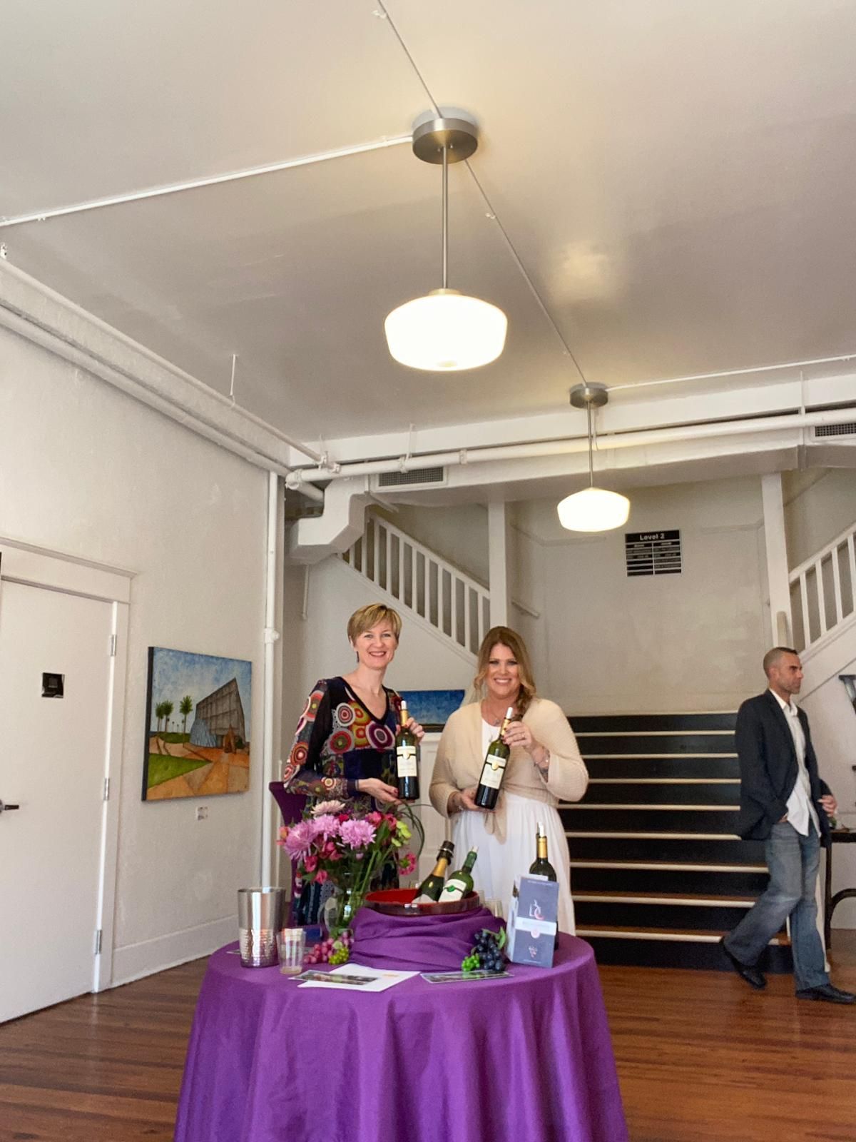 Two women standing next to a table with a purple table cloth holding bottles of wine.