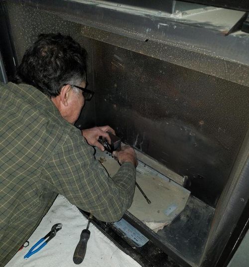 Man in a plaid shirt repairs a fireplace, using tools like pliers and a screwdriver.