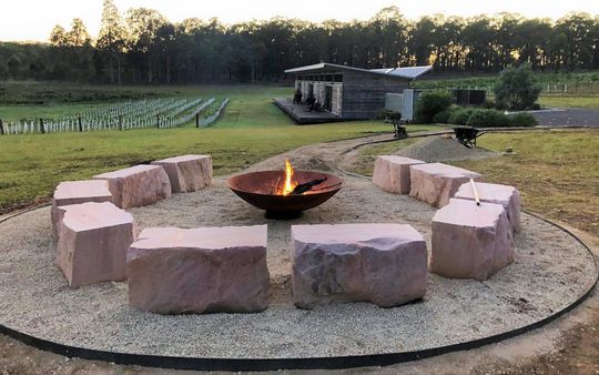 Fire pit with flame, surrounded by stone seating, set on gravel, with vineyard backdrop.