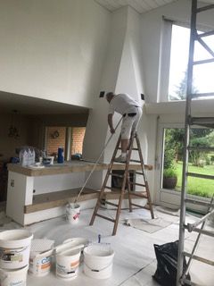 Ein Mann steht auf einer Leiter und streicht eine Wand in einem Wohnzimmer.