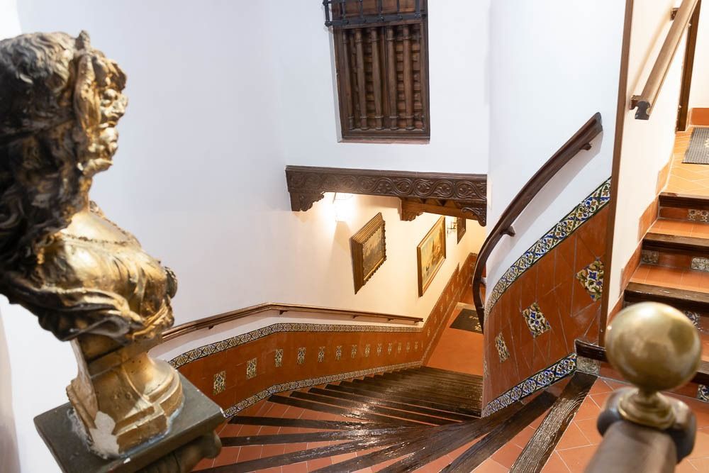 Escalera con paredes y escalones de azulejos. A la izquierda hay un busto. Se aprecian detalles decorativos de madera, obras de arte y una ventana con marco de madera.