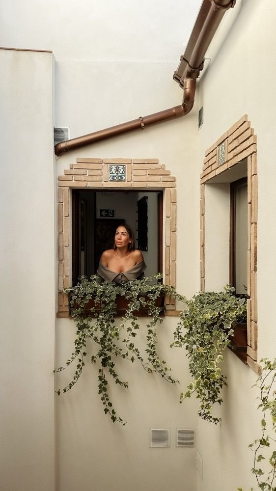 Mujer en una ventana con plantas colgantes, mirando hacia afuera. Paredes color crema, canalón de cobre.