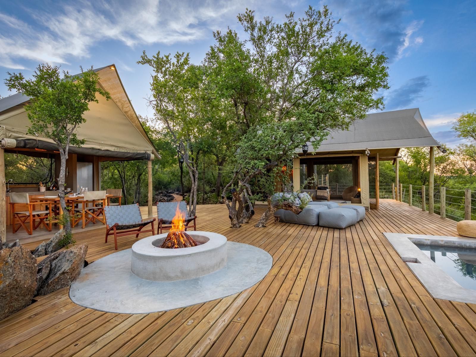 Lodge with wooden deck, fire pit, and lounge area with canvas-covered structures in a lush outdoor setting.