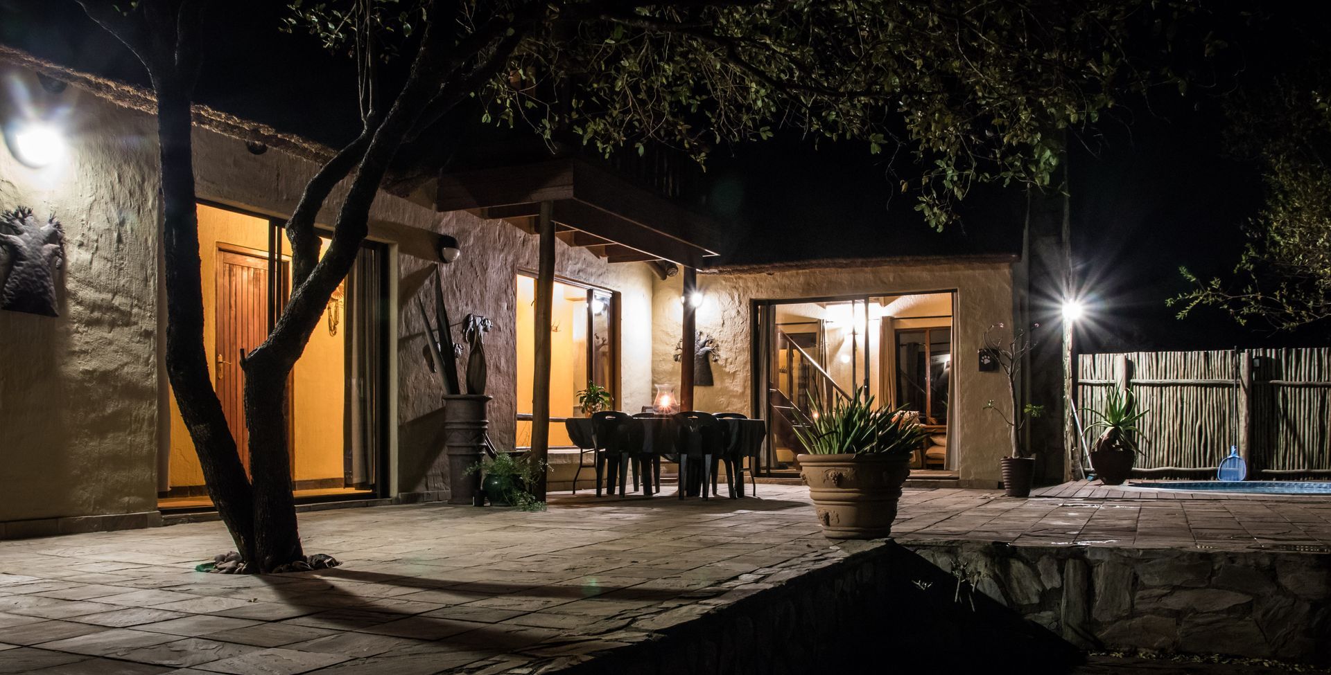 Night view of a Hoedspruit accommodation house with a lit patio and dining table, tree in the foreground, fence to the right 