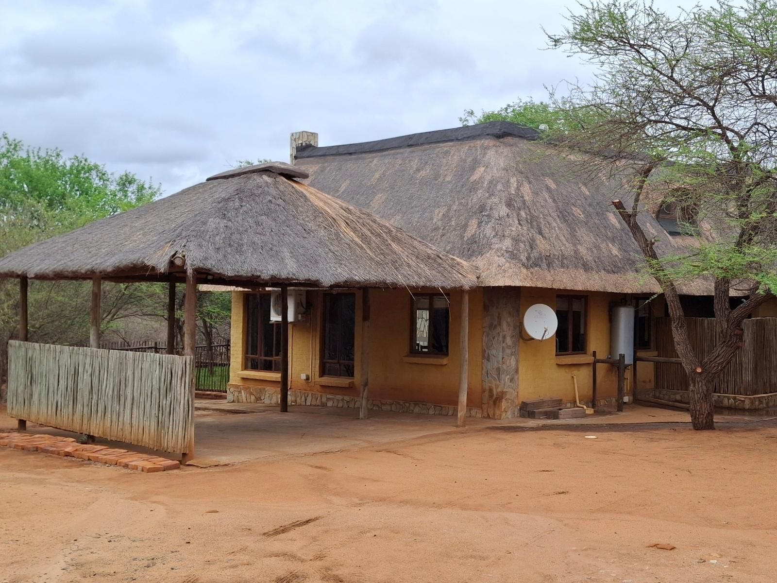 A thatch-roofed, tan-colored bungalow in a dirt yard, with a covered porch and satellite dish.
