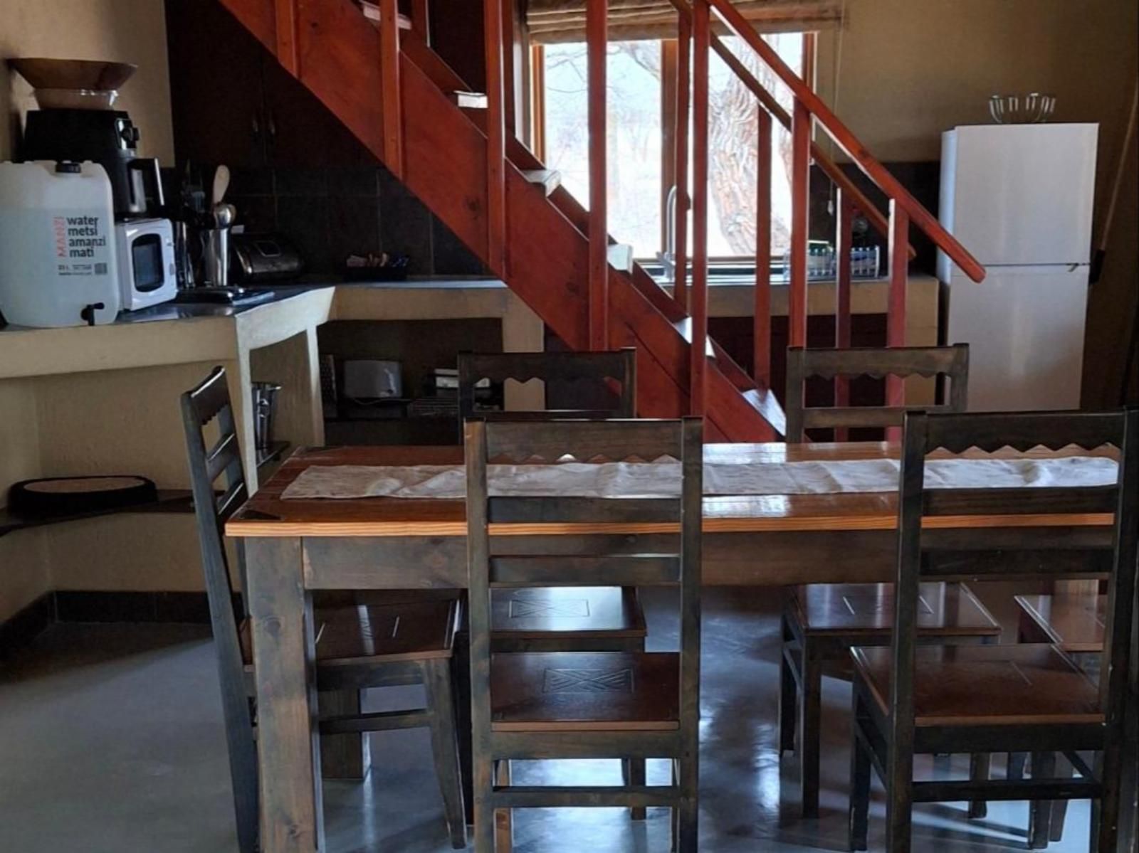 Dining area with a wooden table and chairs, stairs in the background. A refrigerator and kitchen appliances are visible.