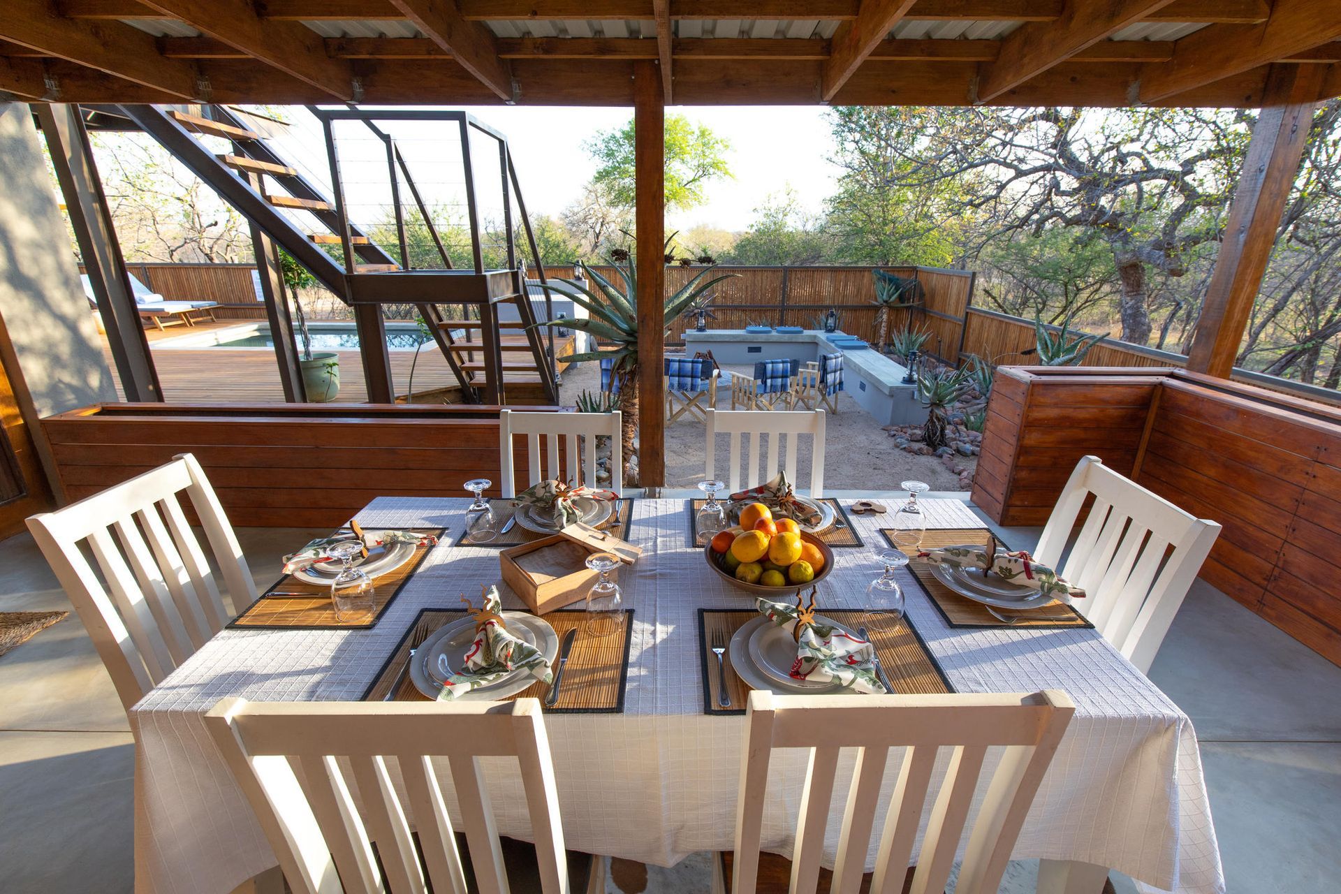 Outdoor dining table set for a meal. White chairs surround a table set with plates, glassware, and fruit in a natural setting.