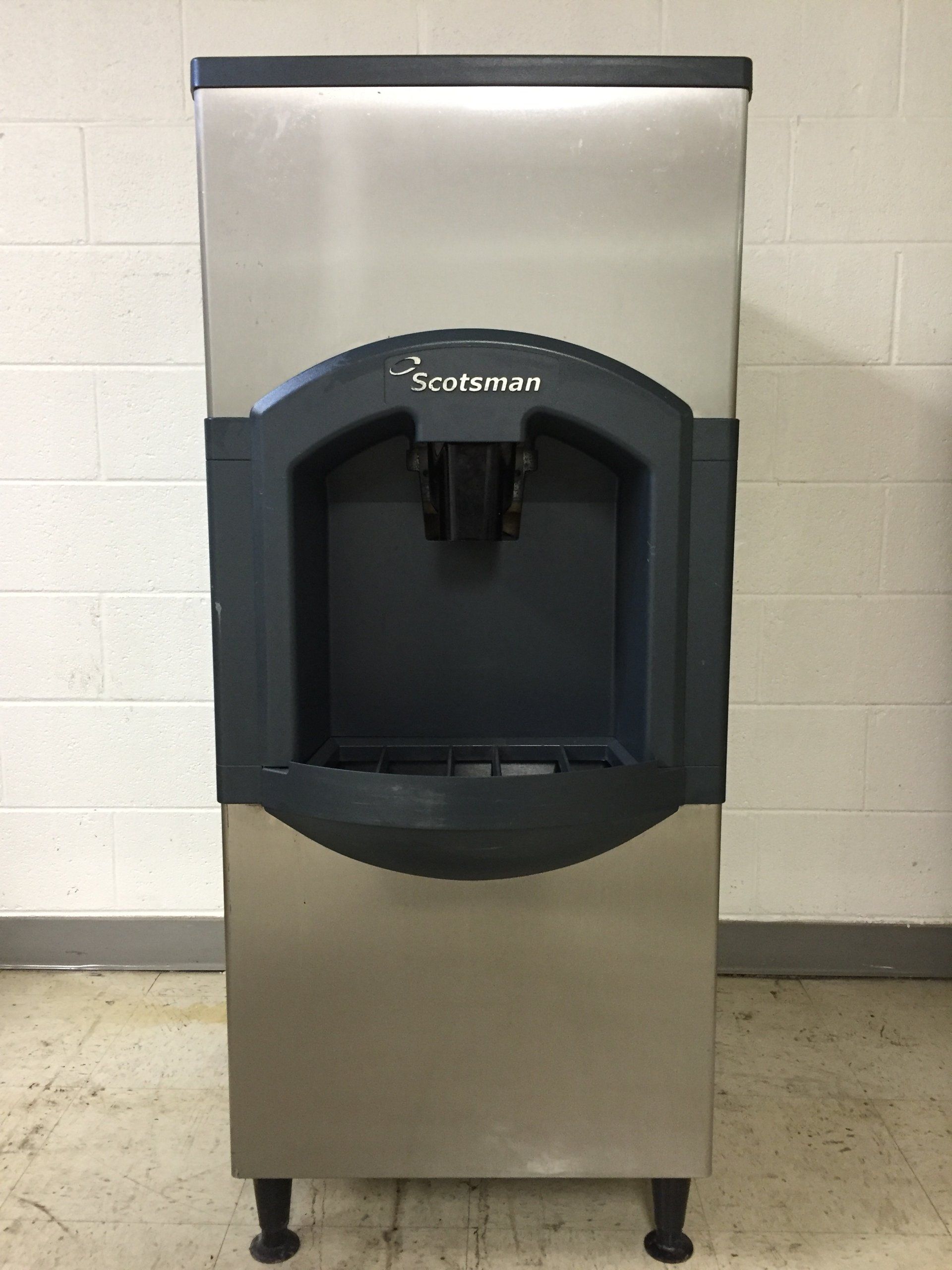 A stainless steel ice machine is sitting in front of a white brick wall.