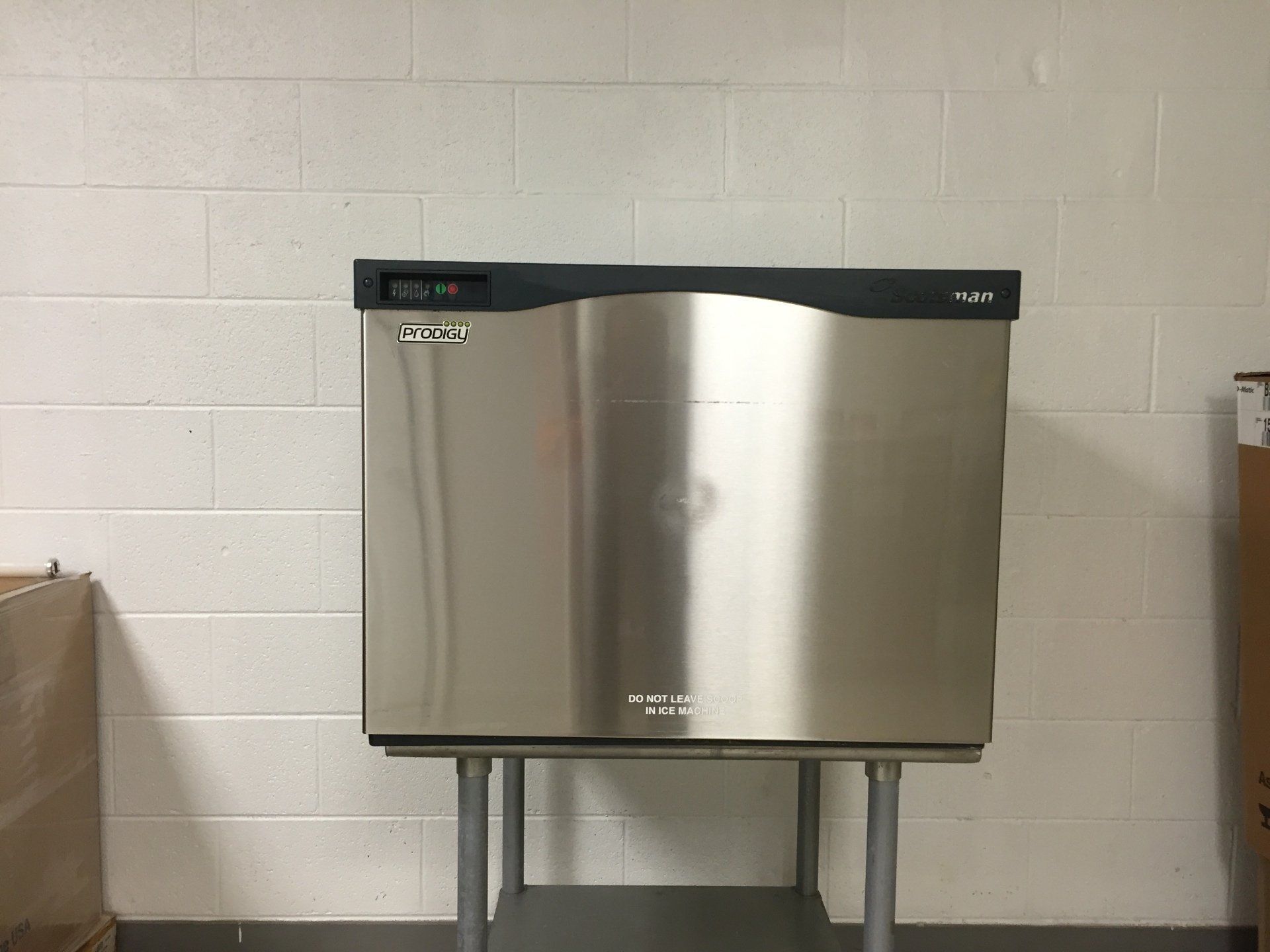 A stainless steel ice makeris sitting on a metal stand in front of a white brick wall.