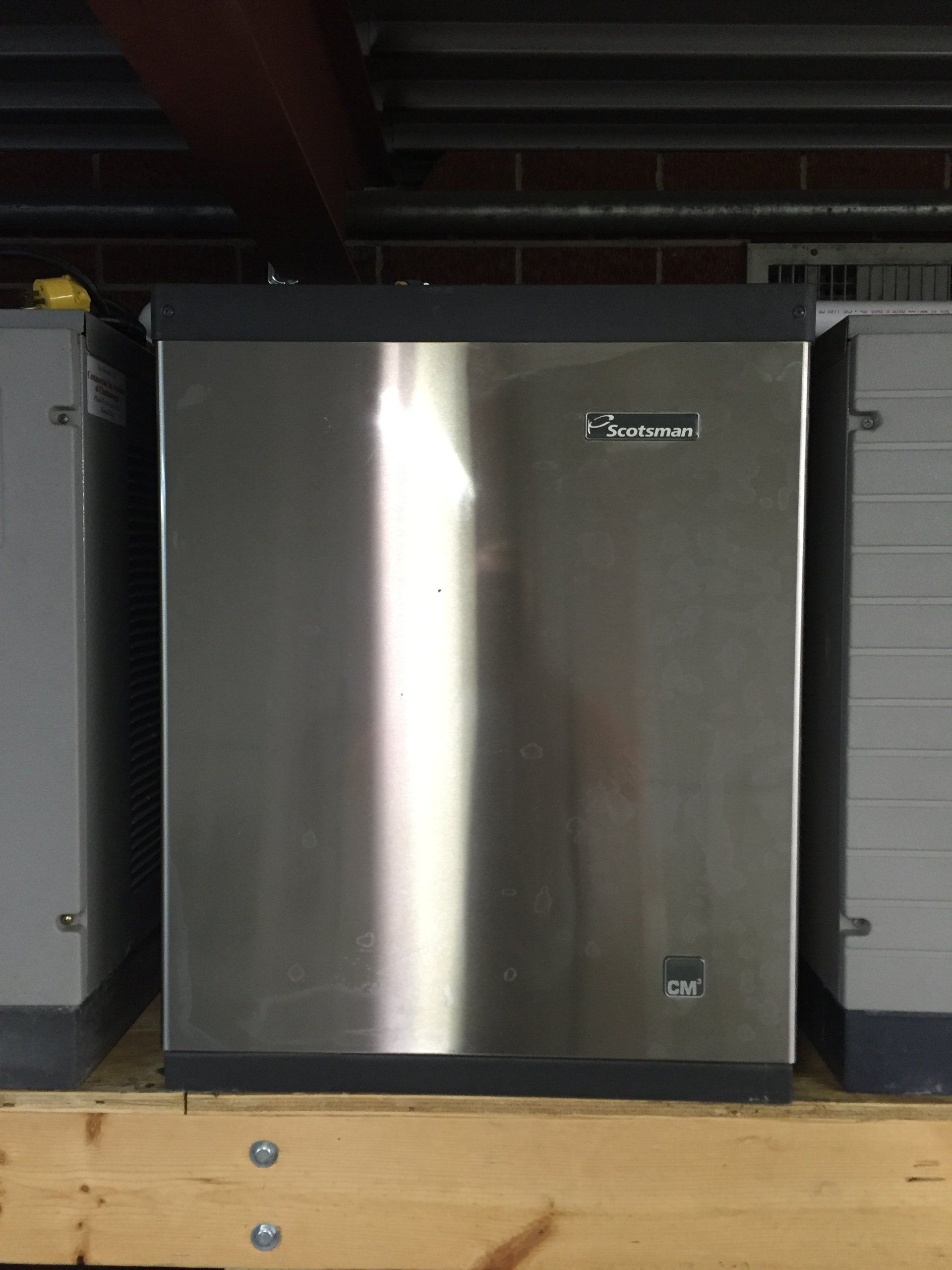 A stainless steel ice maker is sitting on top of a wooden shelf.