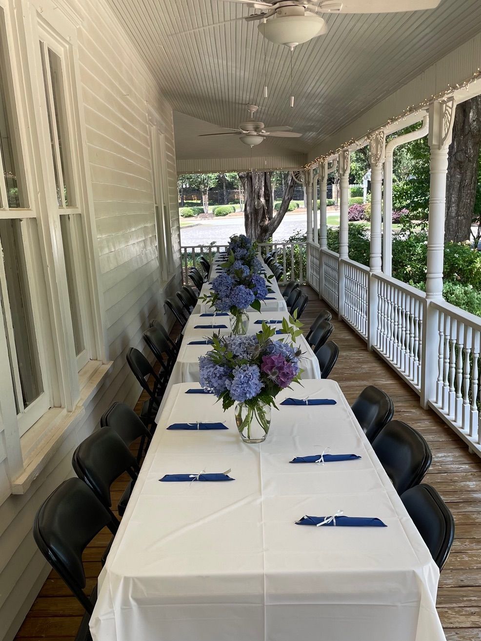Long table set for an event on a porch with floral centerpieces, white tablecloths, and black chairs.