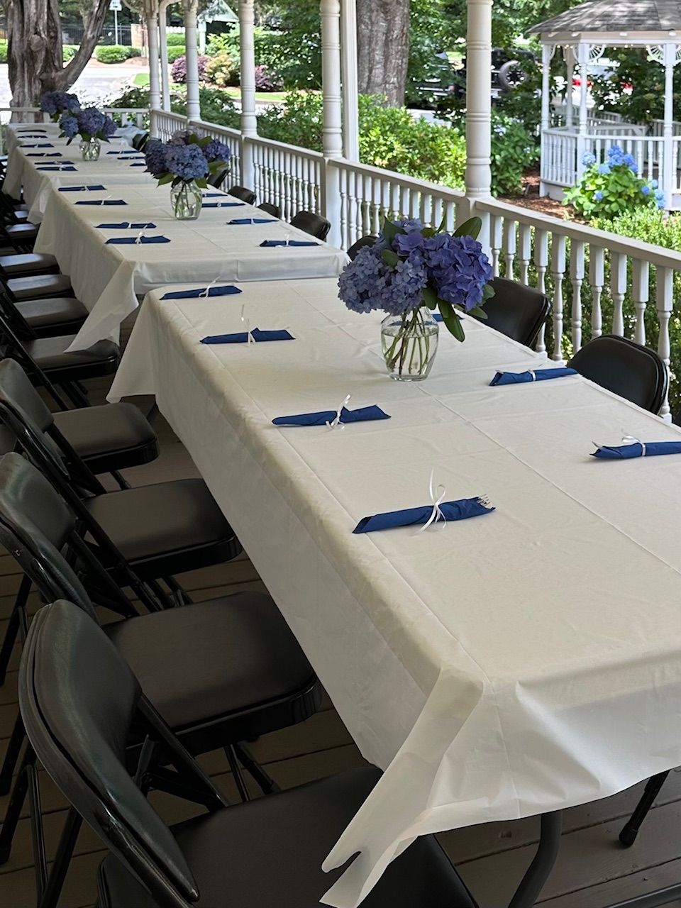 Long tables set for a gathering on a porch, with white tablecloths, blue flowers, and napkins.