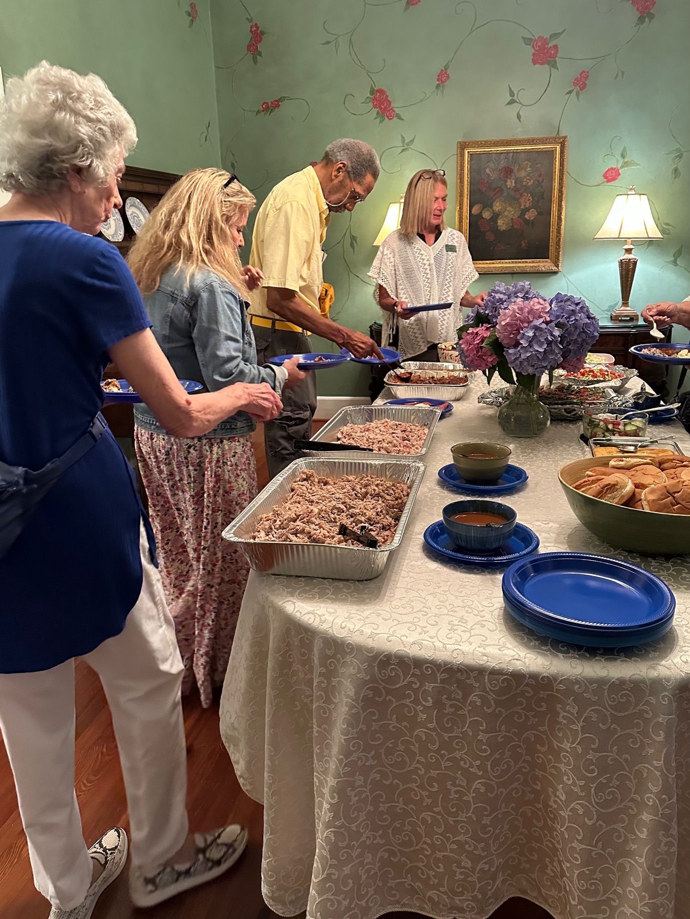 People serving themselves food from a buffet table. Dishes of food, plates, and bowls are arranged on the table.