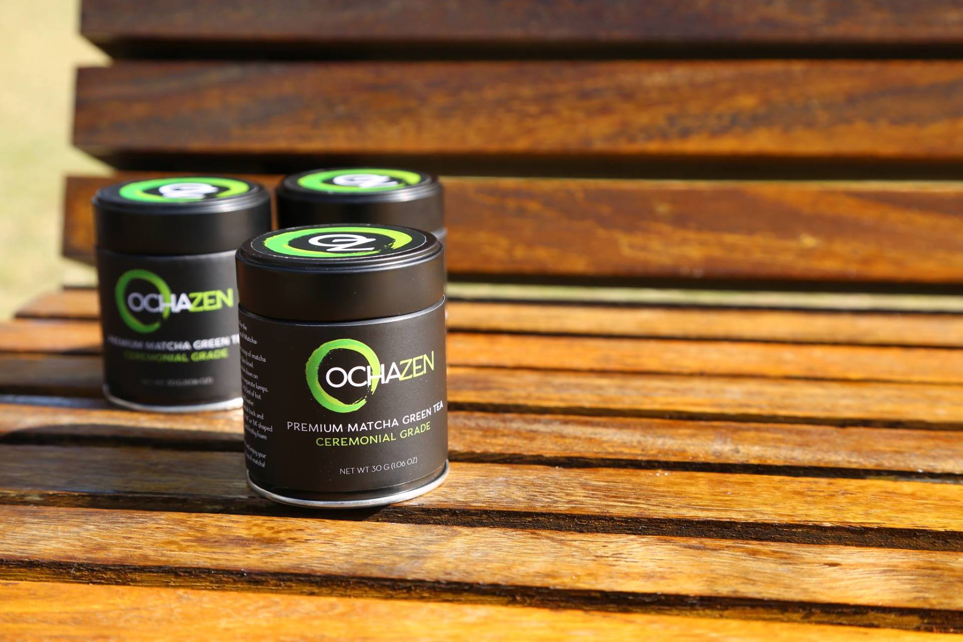 Three black OCRAZEN matcha tea containers on a wooden bench, lit by sunlight.