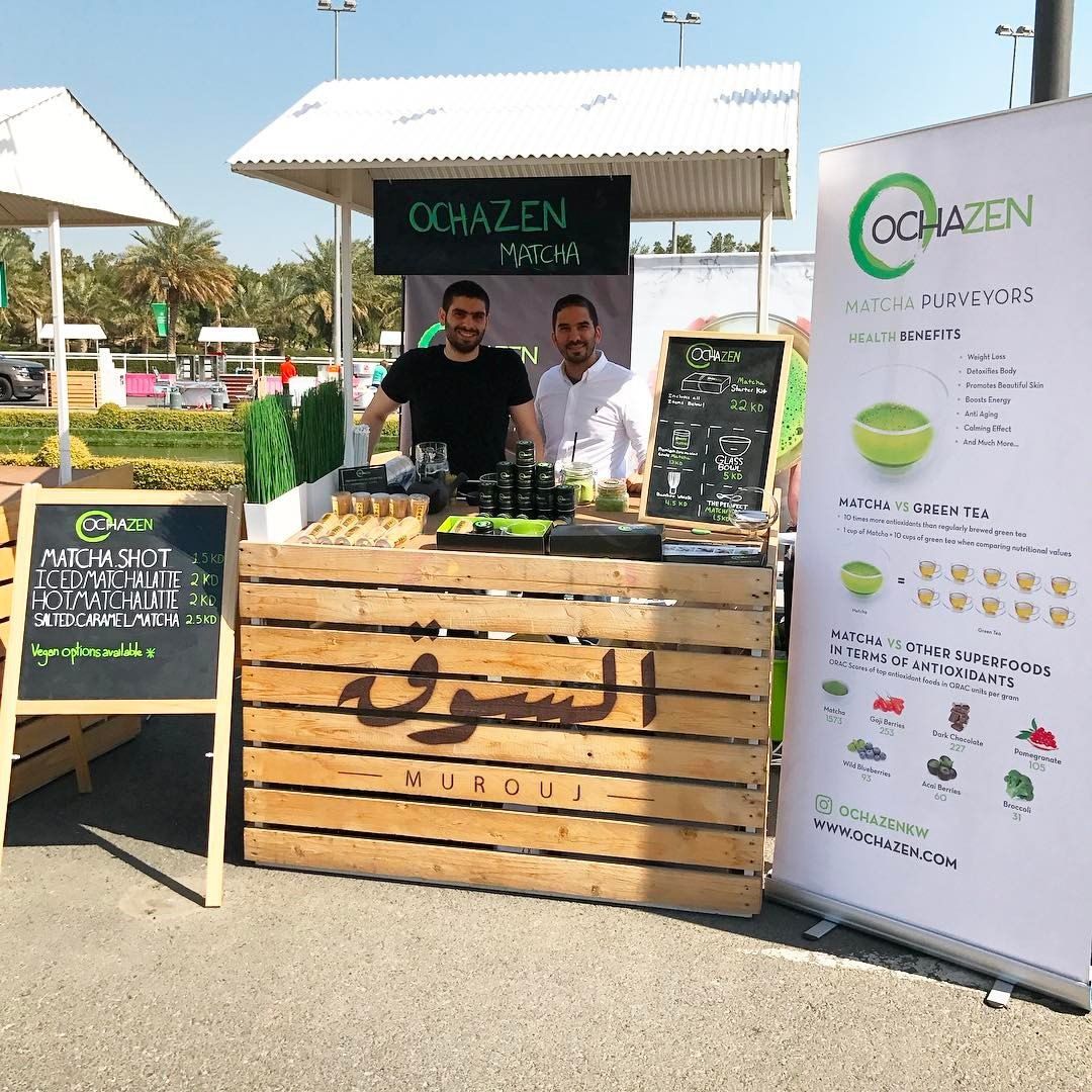 Two men at a food stand called OCHAZEN in a sunny outdoor market, with products displayed.