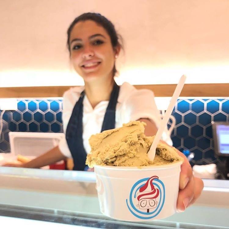 Woman holding out cup of pistachio gelato, smiling behind counter with blue honeycomb tiles.