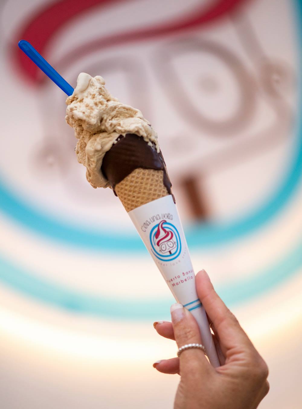Hand holding an ice cream cone with two scoops of ice cream, partially dipped in chocolate, and a blue spoon.